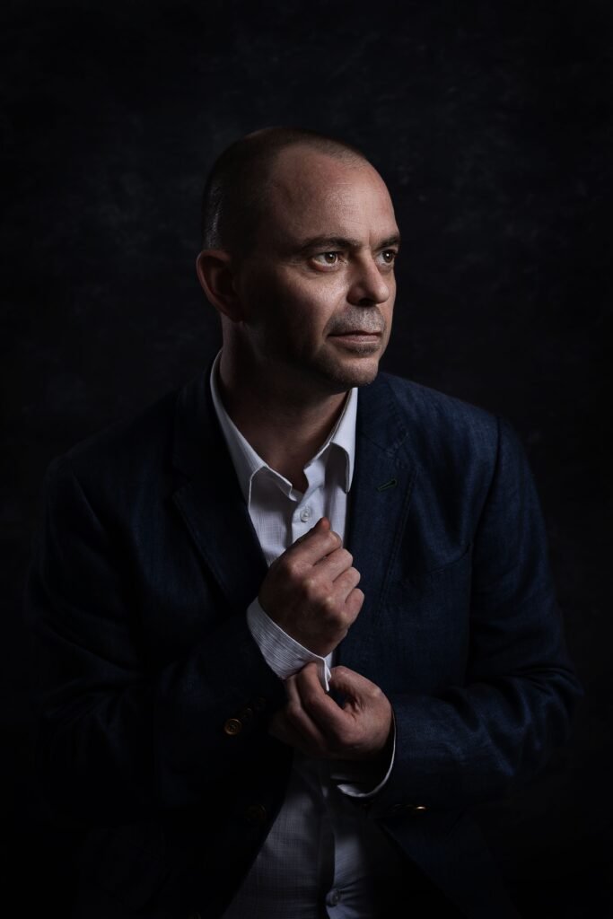Moody portrait of an adult man in a black suit and white shirt against a dark background.